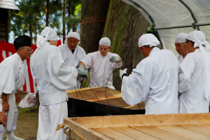 写真：白装束の男性が数人がかりで大きな網に乗せた油揚げを油の鍋の中から持ち上げ、揚げ具合を確認しています