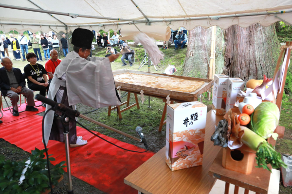 写真：神主が大きな油揚げのお祓いをしているところ。祭壇にはニンジン・キャベツ・大根などの野菜や日本酒などが奉納されています