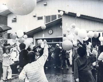 コミセン入口に集まった地域住民でバルーンリリースをする様子を撮影した白黒写真