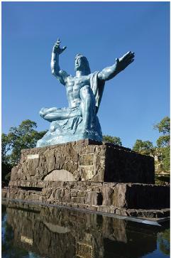 写真：長崎の平和記念像。像は、右手を垂直に掲げ、左手は水平に伸ばし、軽く目を閉じ柔和な顔をした男性のようです。