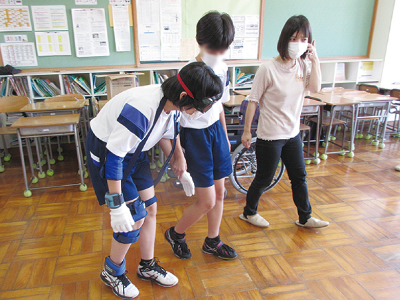 体の動きを妨げる装置を付けた中学生。もう一人の中学生と先生が寄り添っています