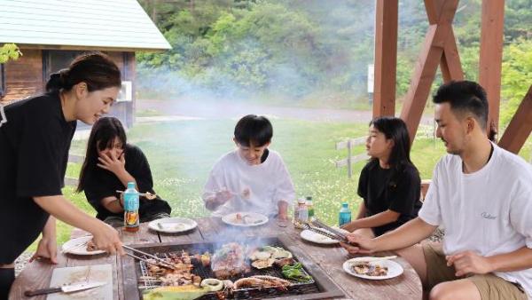 写真：談笑しながらバーベキューを楽しむ5人の家族。お母さんが立ち上がってお肉の焼け具合を見ています。
