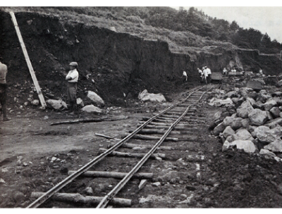 ダム建設当時の写真（モノクロ）。土砂を運ぶためのトロッコの線路があり、その脇には工事で出てきた人間の頭以上の大きさの石が積みあがっています。