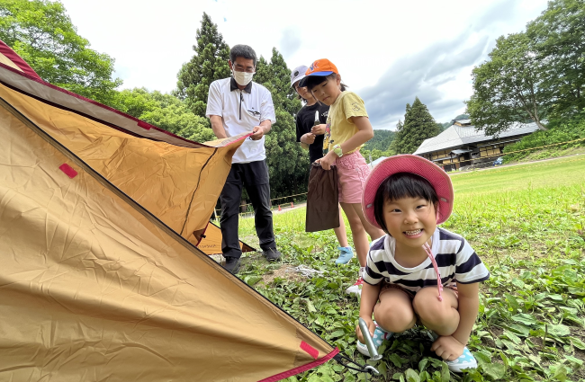 写真：大きなテントを張る男性と3人の子ども。子どもの1人がワクワクした顔でカメラを見ています。