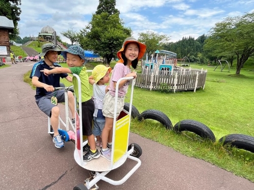 写真：おもしろ自転車の1つ。お父さんの漕ぐ自転車の前方に、人が立って乗れるカゴがついていて、3人の子どもが載っています。