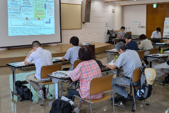 写真：講師がスクリーンに映し出した資料の説明を聞く訓練参加者