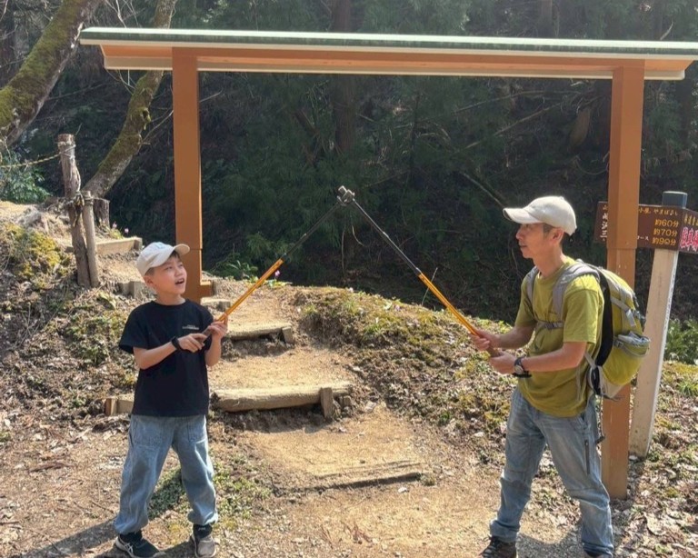写真：登山口でお父さんと子どもがストックの先を合わせています