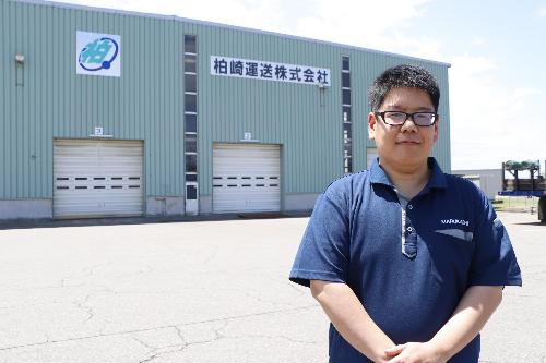 写真：会社の大きな倉庫前に立っている橋本さん