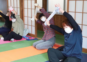 写真：タオルの両端を持った手を頭の上に上げ、体の脇を伸ばす体操をする参加者