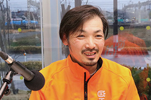 蛍光オレンジの森林ウエアを着た柏崎地域森林組合の従業員、中村貴洋さんがマイクに向かって話しています。