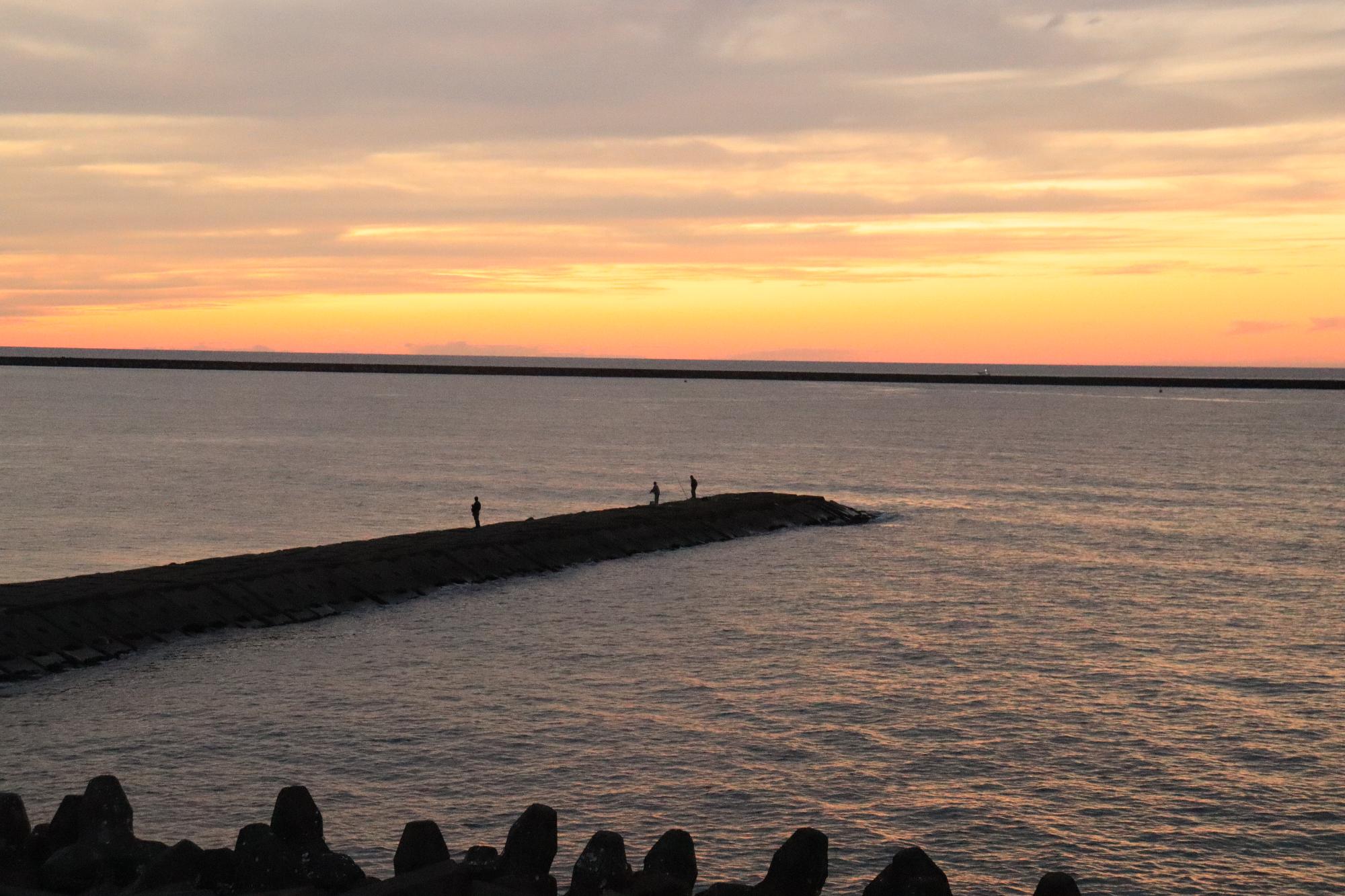 夕日に染まる日本海と空。釣り人のいる防波堤とテトラポットが映っています