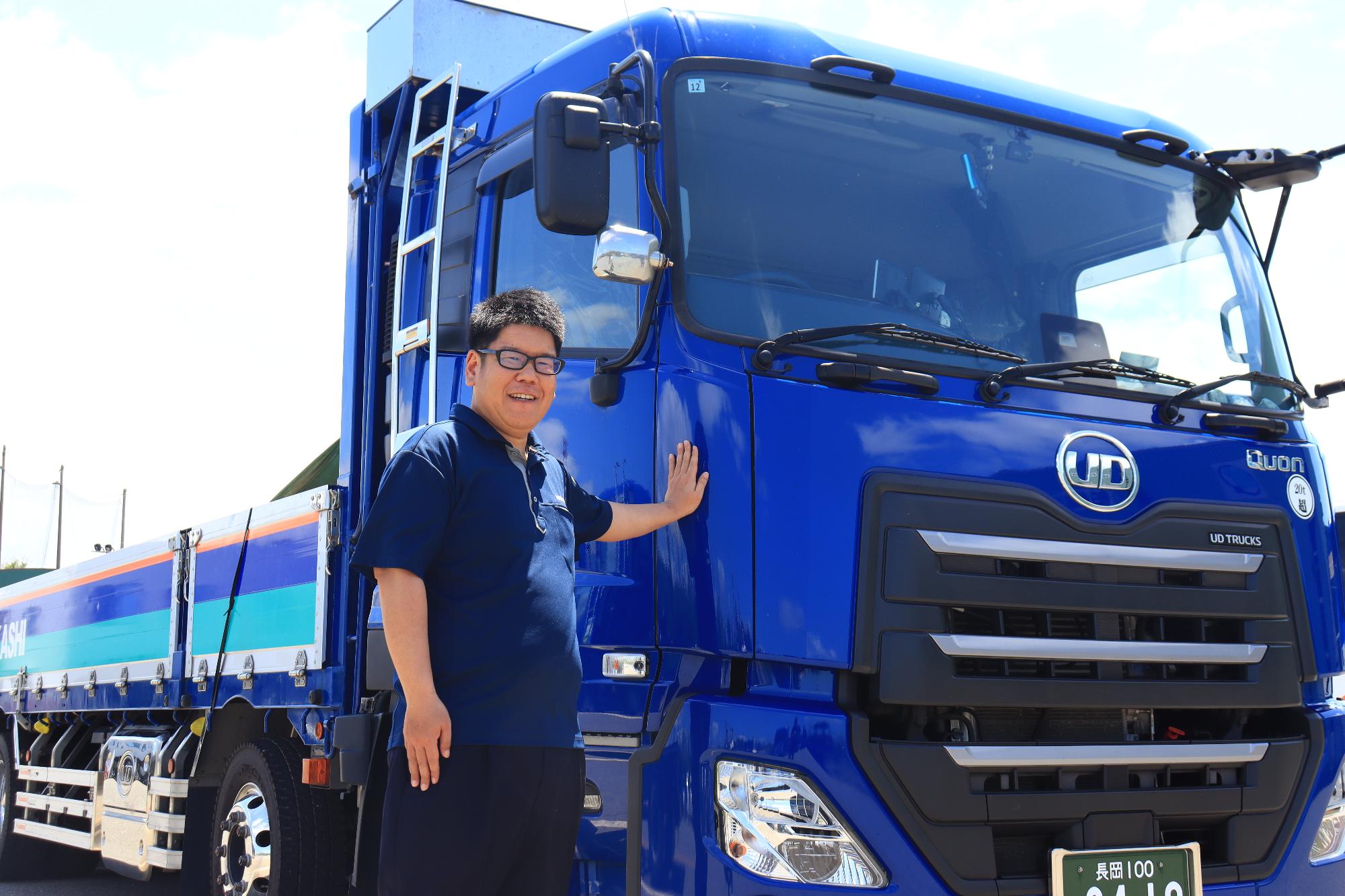 写真：青色の車体のトラックに左手を当てポーズをとる橋本さん