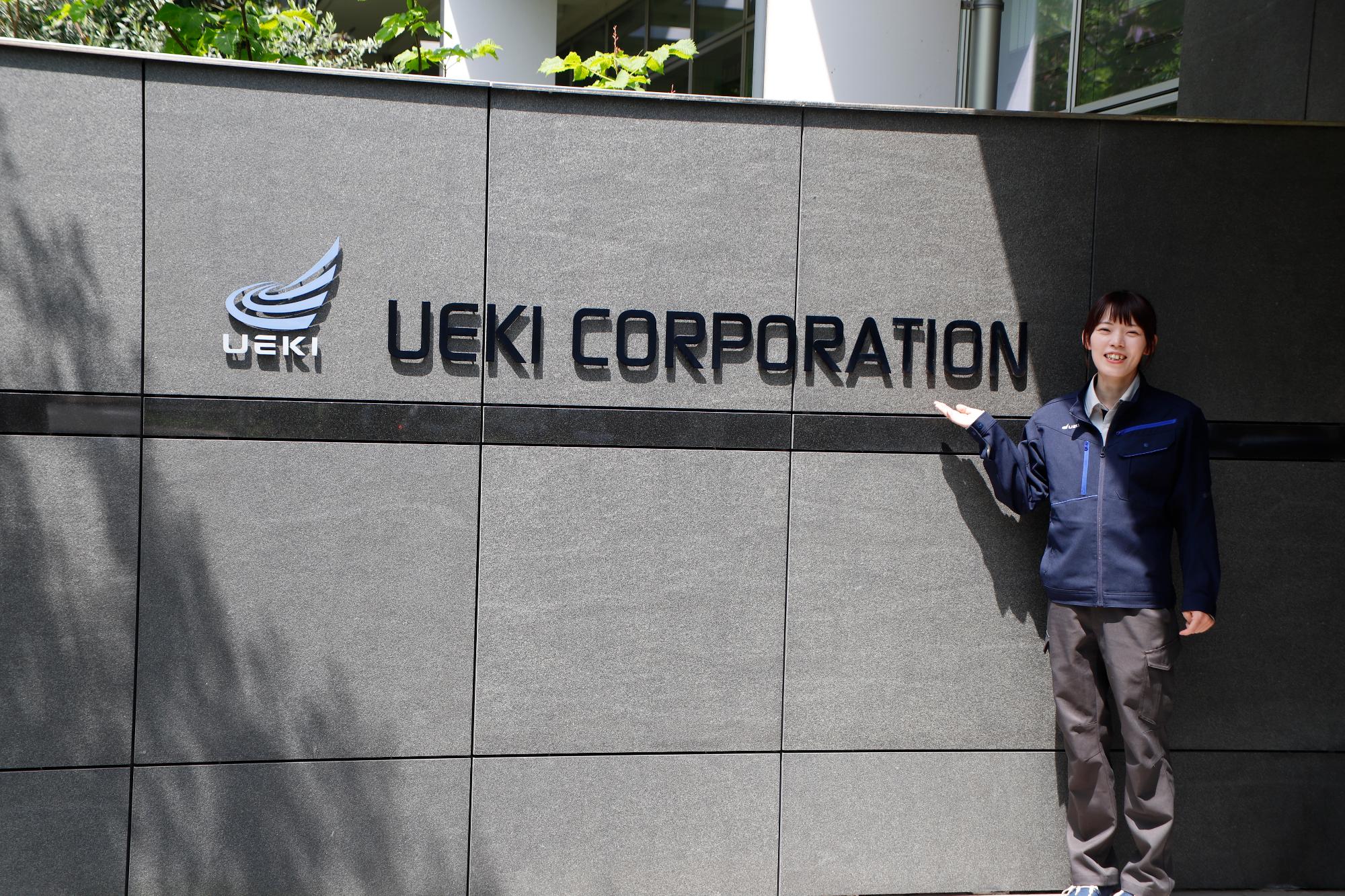 写真：株式会社植木組の本社前で、社名に手を添えたポーズをとる橘さん