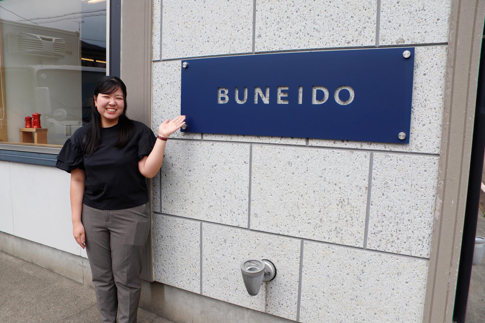 写真：「BUNEIDO」とローマ字でかかれた会社の看板の前で、にこやかな笑顔を浮かべる田川さん